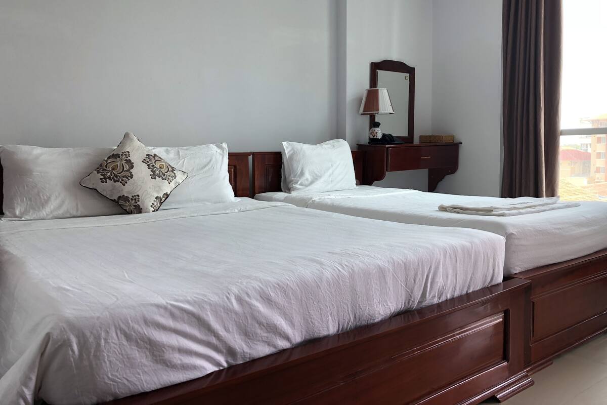 A hotel room with two beds, a wooden headboard, a nightstand with a lamp, and a window with curtains.
