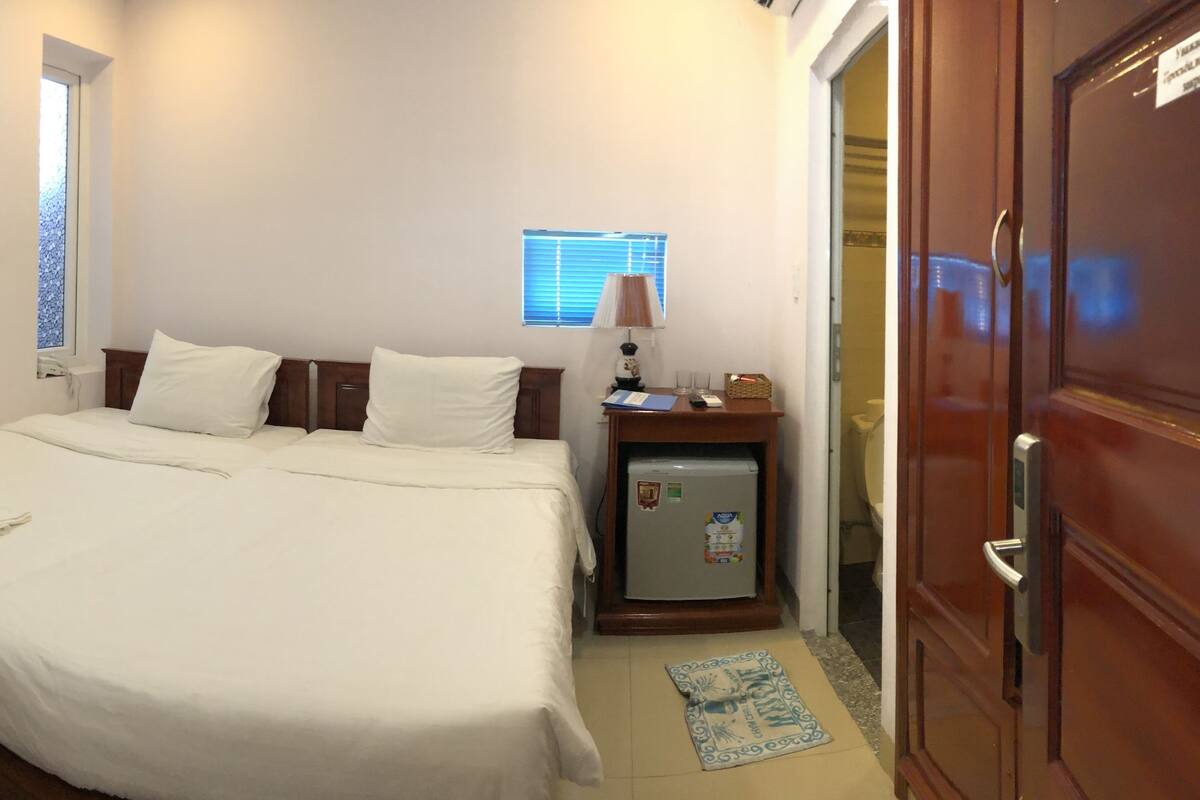 A neatly made bed with white linens, a small bedside table with a lamp, and a refrigerator.