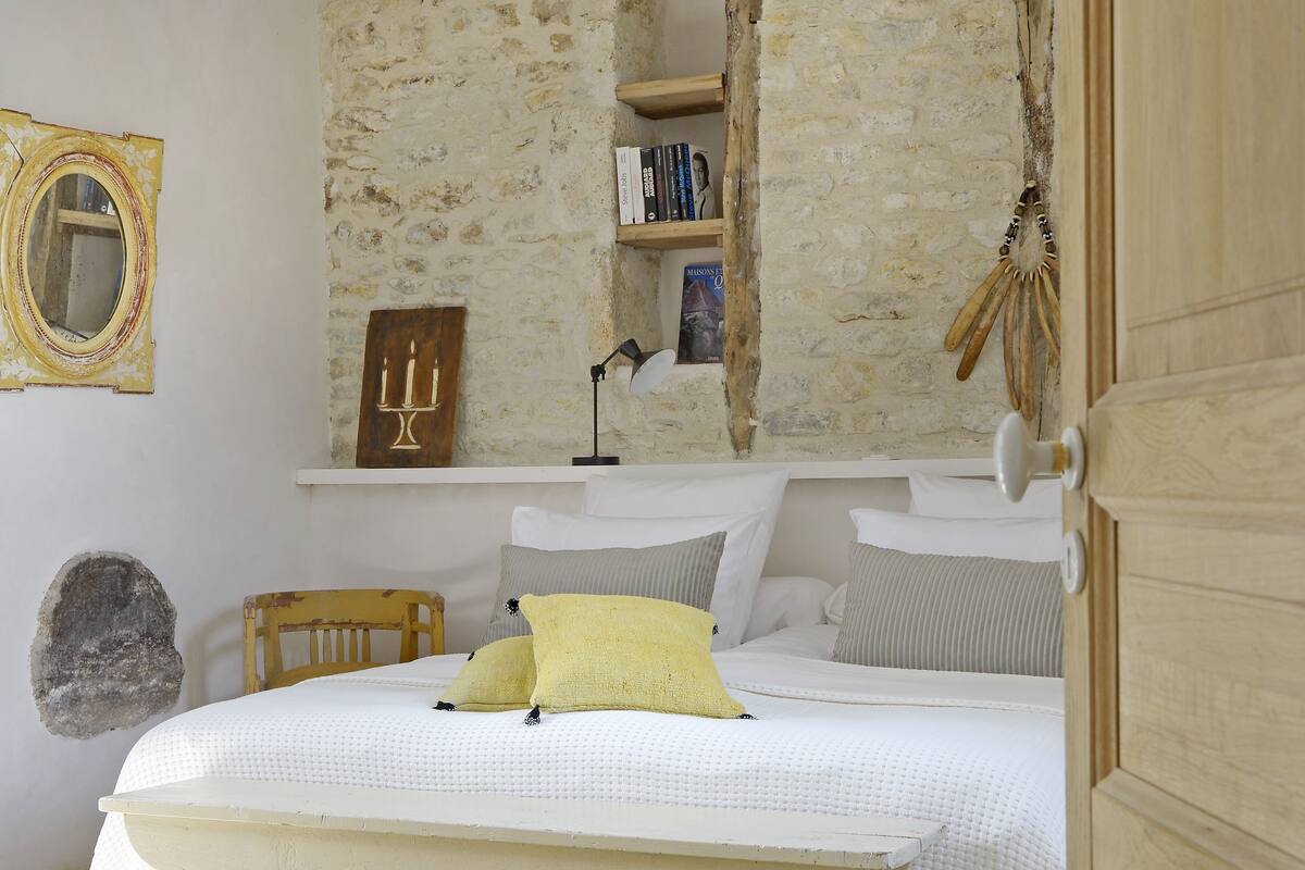 A bedroom with a bed, a wooden headboard, a bedside table, a lamp, a mirror, and a stone wall.