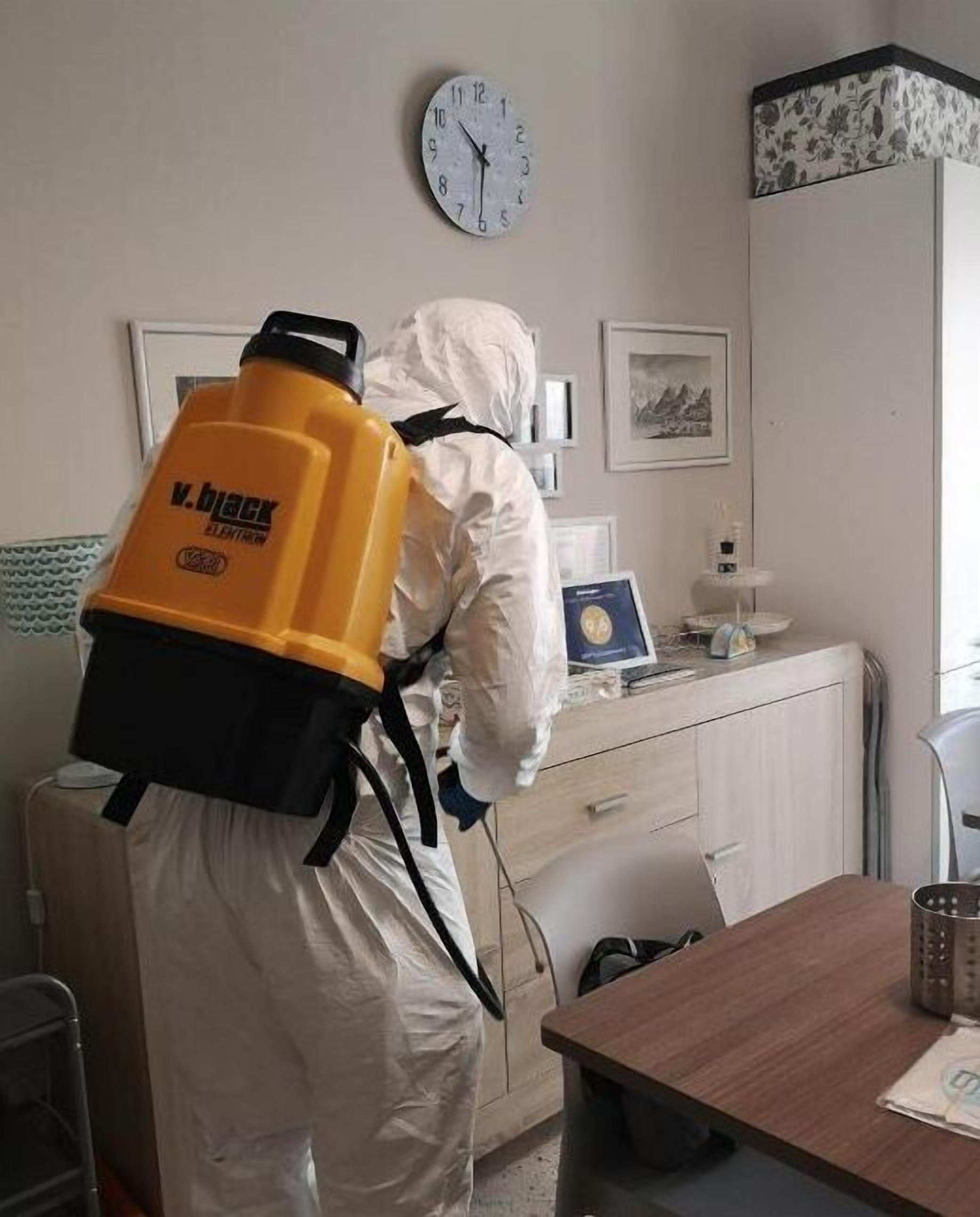 Una persona amb un vestit protector blanc i una motxilla groga, dempeus en una habitació que té un rellotge, una fotografia amb marc i un escriptori amb un ordinador portàtil.