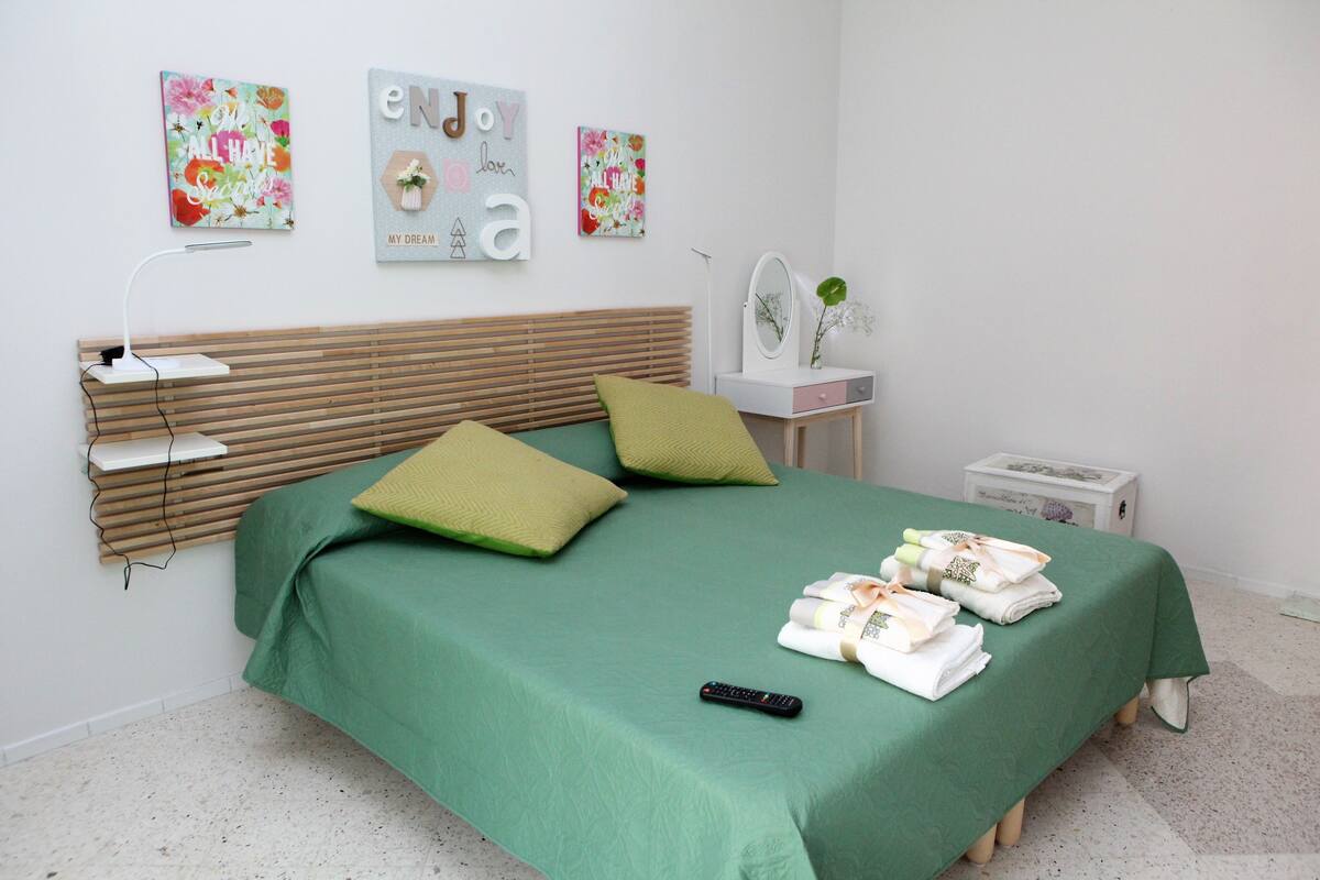 Ein ordentlich gemachtes Bett mit einer grünen Tagesdecke, zwei Kissen und einer Fernbedienung. An den Wänden befinden sich Kunstwerke und ein kleiner Nachttisch mit einem Spiegel und einer Blumenvase.