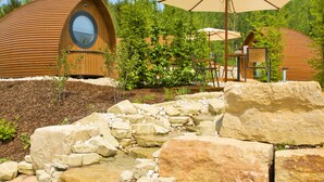 Una cabaña moderna de estilo rústico con ventana circular, rodeada de exuberante vegetación y una cascada pequeña.