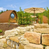 Una cabaña moderna de estilo rústico con ventana circular, rodeada de exuberante vegetación y una cascada pequeña.