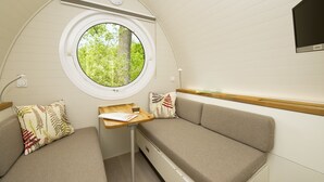 Un interior compacto y bien iluminado, con un ventanal redondo que ofrece vistas a la vegetación, una pequeña mesa de madera y dos zonas de estar con cojines.