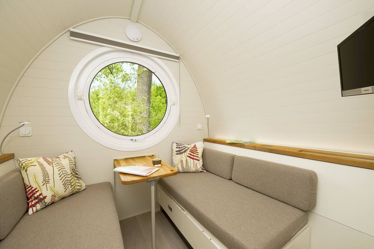 Un intérieur compact et bien éclairé, doté d’une fenêtre ronde offrant une vue sur la verdure environnante, d’une petite table en bois et de deux coins salon agrémentés de coussins.