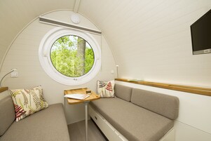Un interior compacto y bien iluminado, con un ventanal redondo que ofrece vistas a la vegetación, una pequeña mesa de madera y dos zonas de estar con cojines.