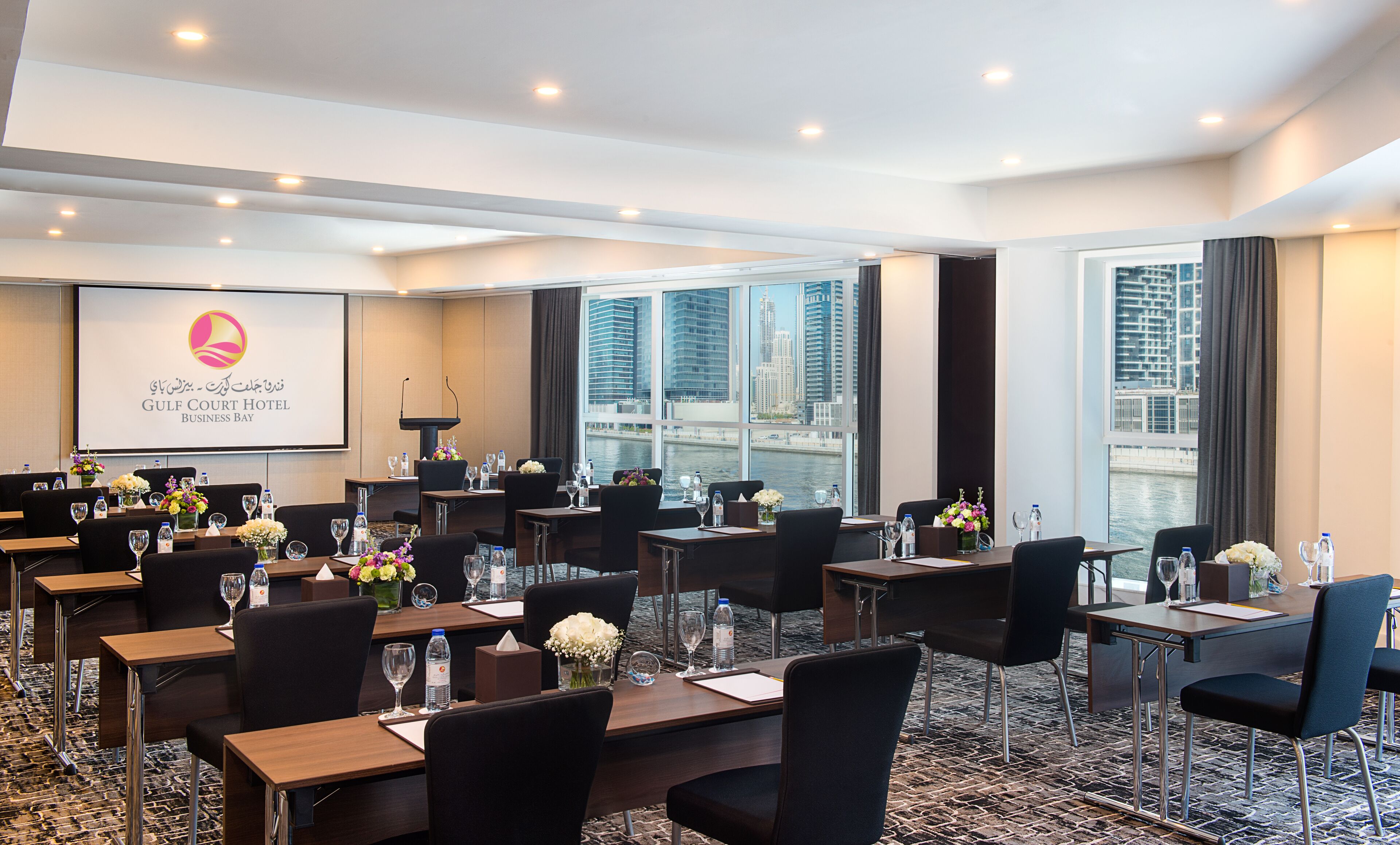 Une salle de conférence à l’hôtel Gulf Court Business Bay, équipée de plusieurs tables, de chaises et d’un grand écran affichant le logo de l’hôtel.