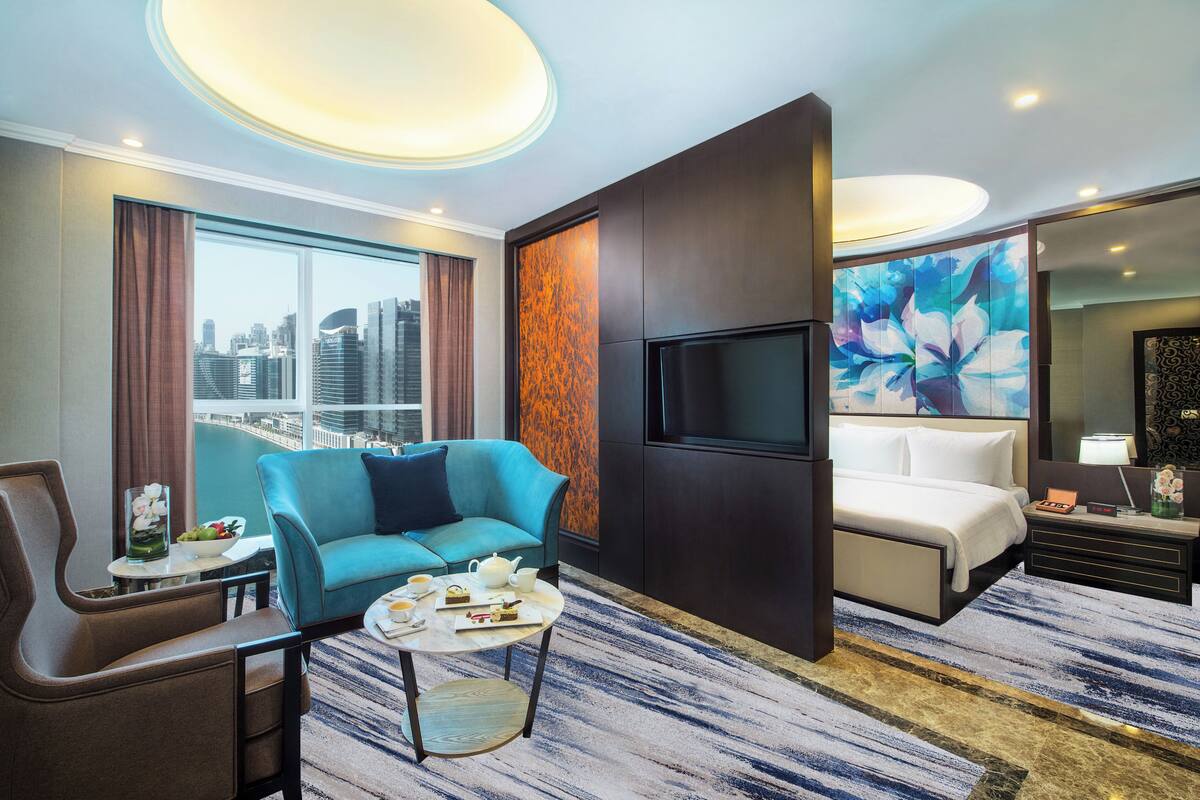A modern hotel room with a city view, a sofa, a bed, a TV, and a coffee table with snacks.