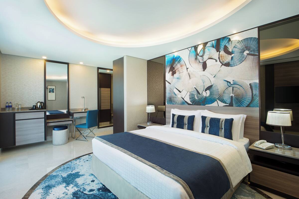 A modern hotel room with a large bed, a desk, a chair, a bathroom, and a large abstract painting on the wall.