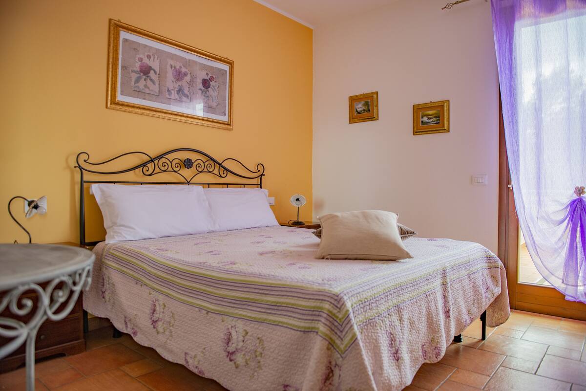 Een slaapkamer met een bed, nachtkastje, stoel en een raam met gordijnen.