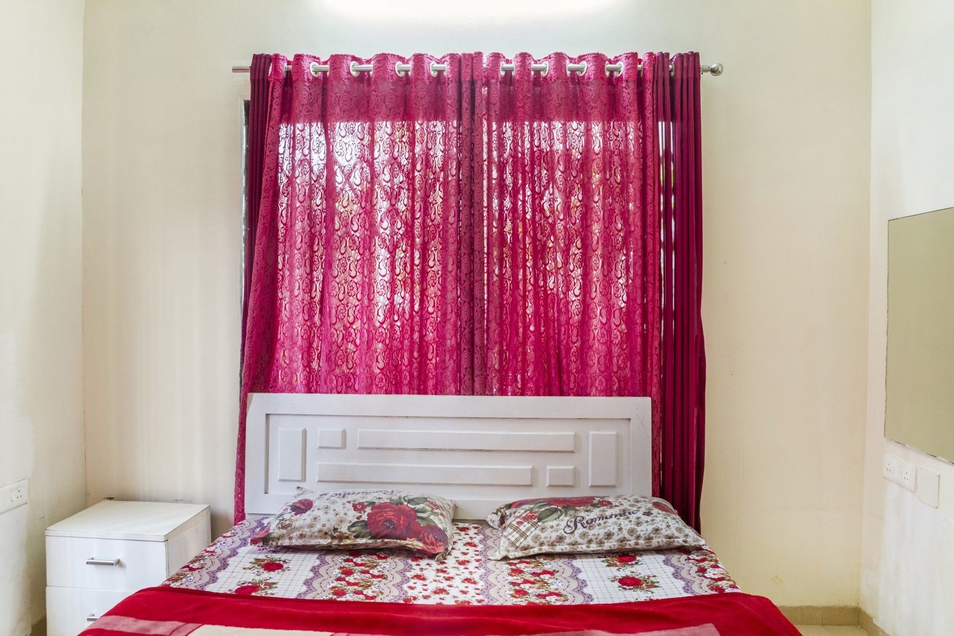 A bedroom with a bed, two pillows, a nightstand, and a window with red curtains.