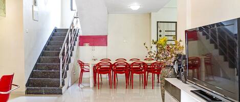 A modern living room with a staircase, a dining area with red chairs, and a flat-screen TV on a cabinet.