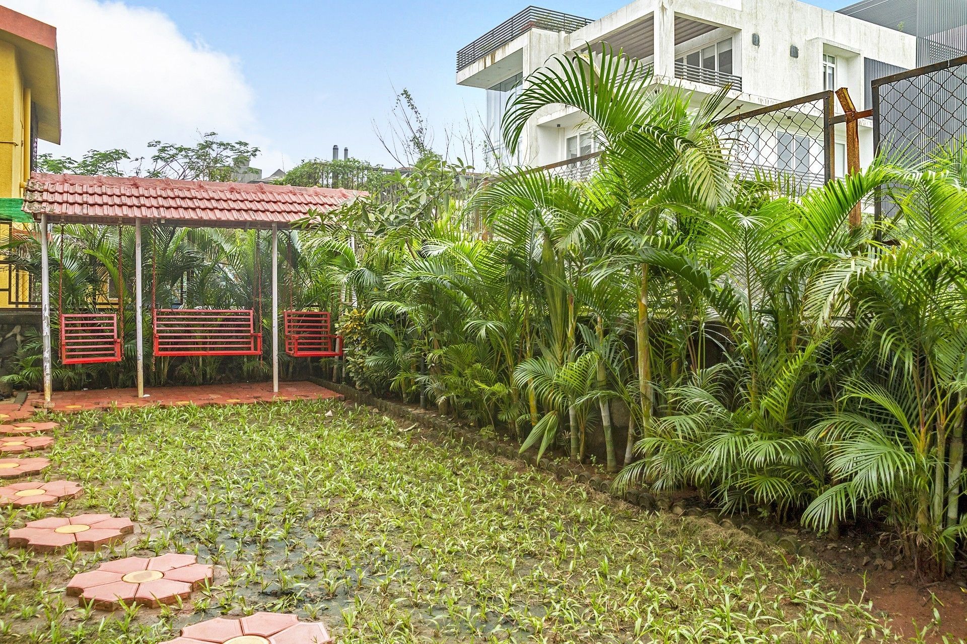 Un jardin avec une balançoire, une zone pavée où sont plantées de jeunes plantes, et un bâtiment à la façade blanche et doté de balcons.