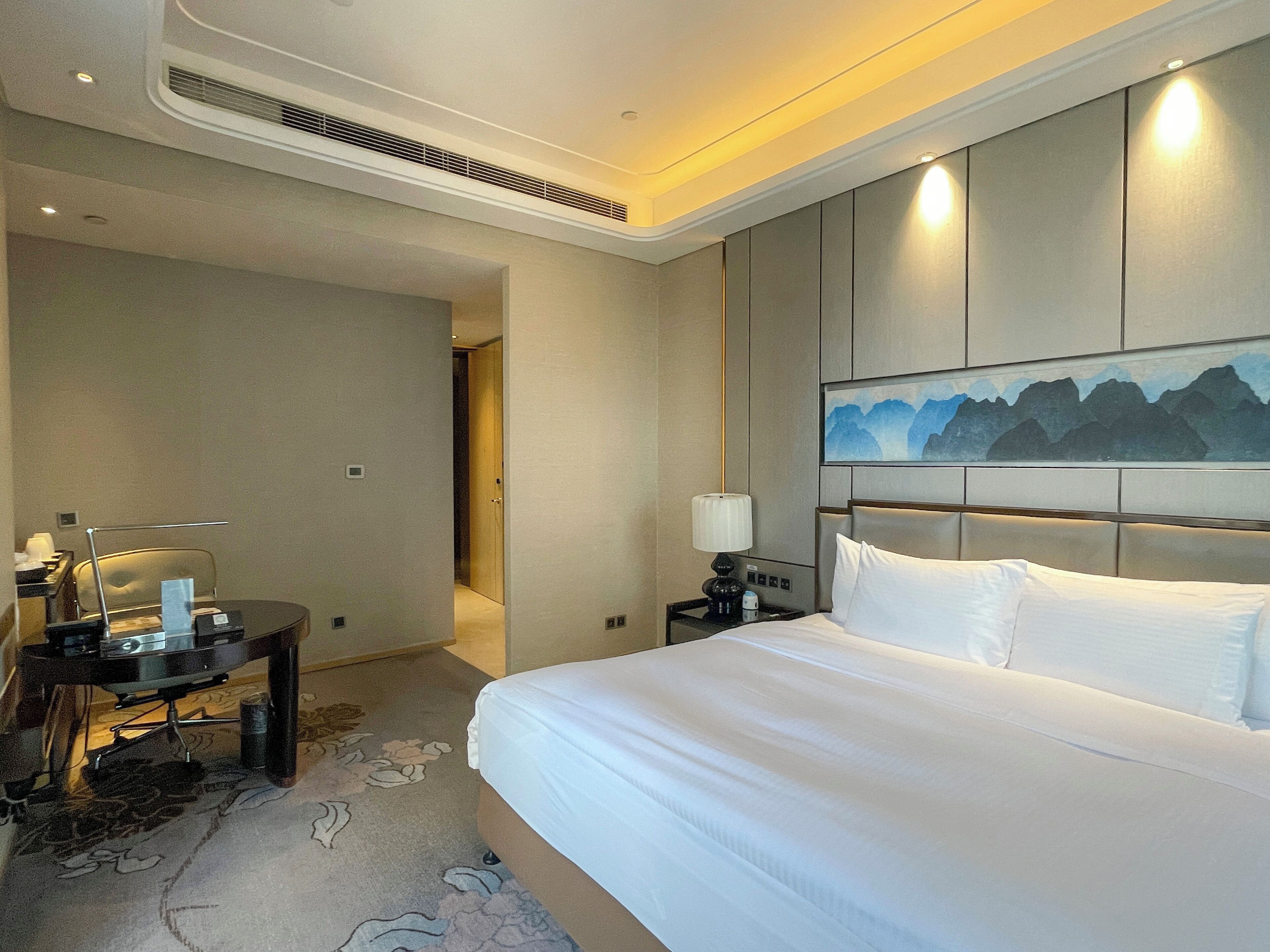 A hotel room with a large bed, a desk with a chair, a lamp, and a painting on the wall.