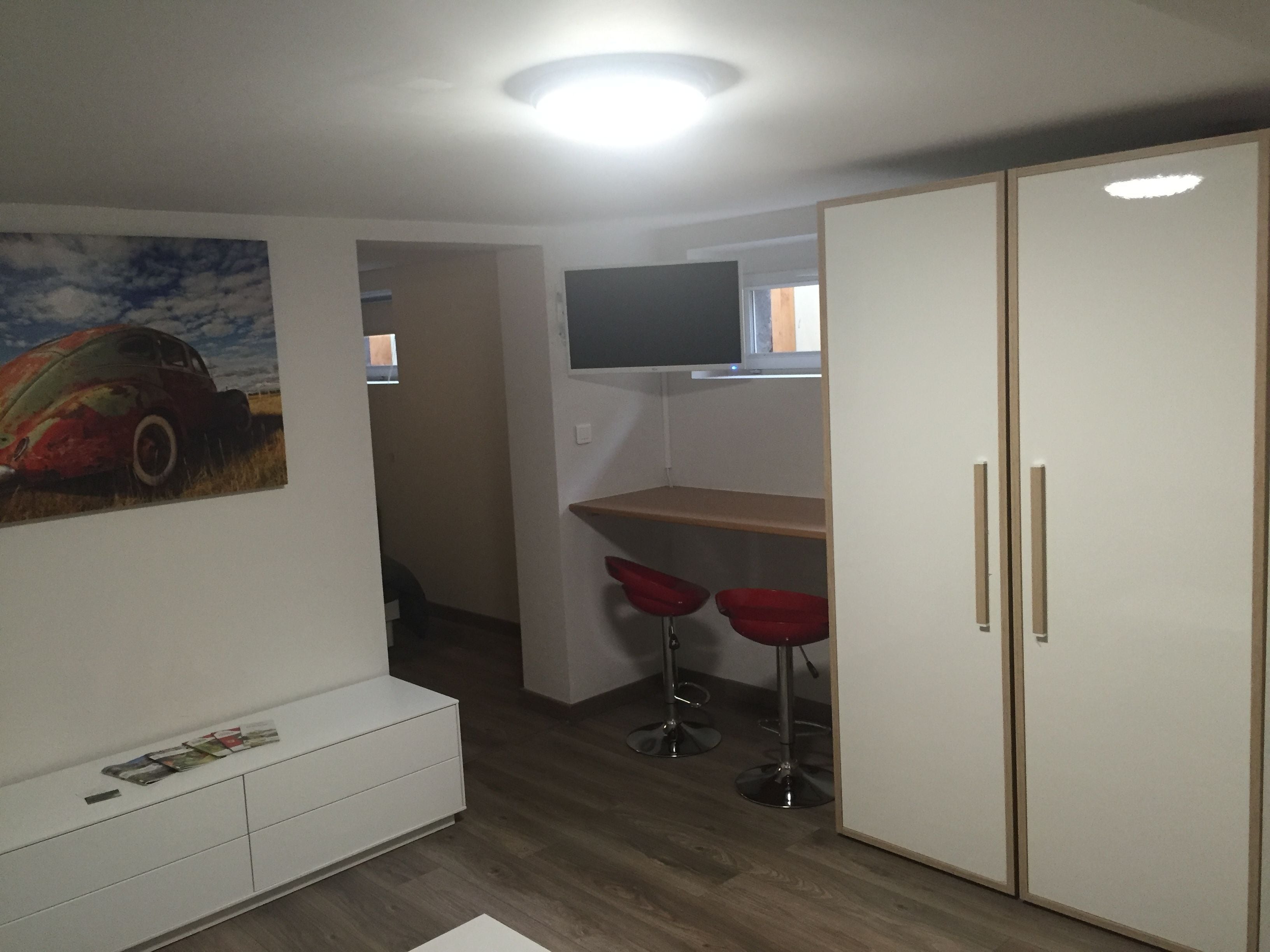 A room with a white wardrobe, a TV mounted on the wall, and two red bar stools.