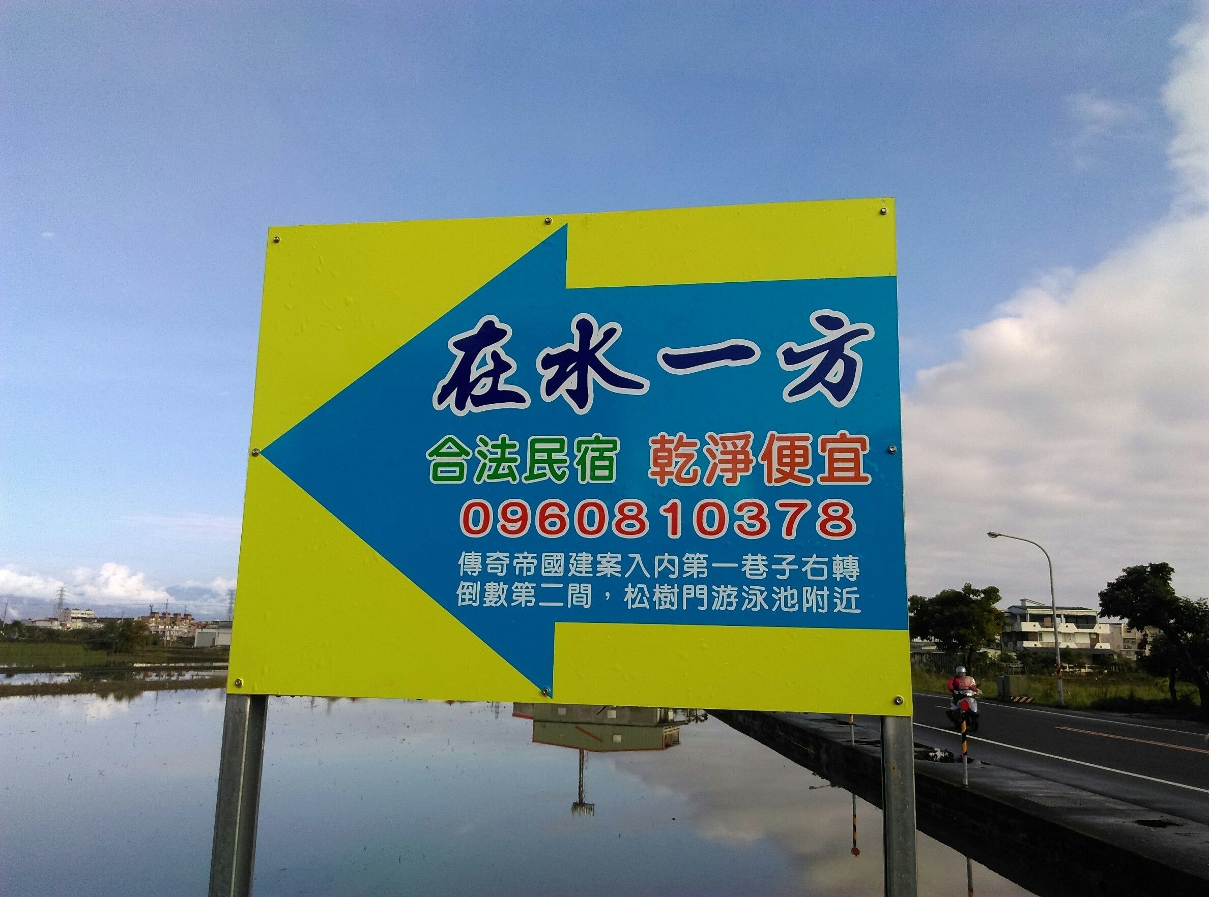 A sign with text and a phone number, placed near a body of water with a road and a cyclist in the background.