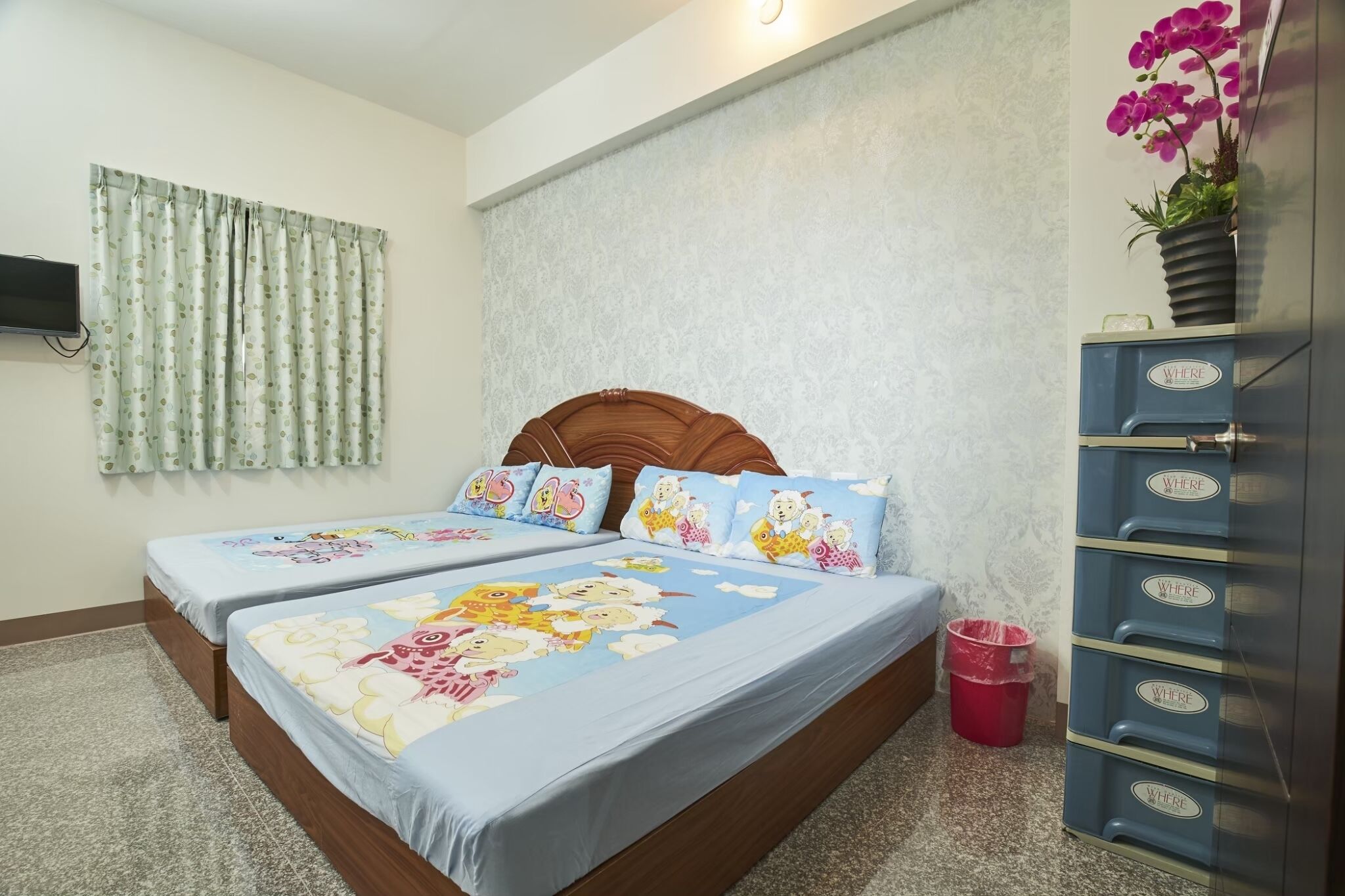 Un dormitorio con dos camas, un televisor montado en la pared, una cortina estampada, una planta decorativa y un mueble de almacenamiento con etiquetas que dicen 'DONDE'.