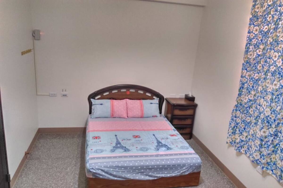 Camera da letto con un letto, una testiera in legno, un comodino, una tenda floreale e una presa elettrica.