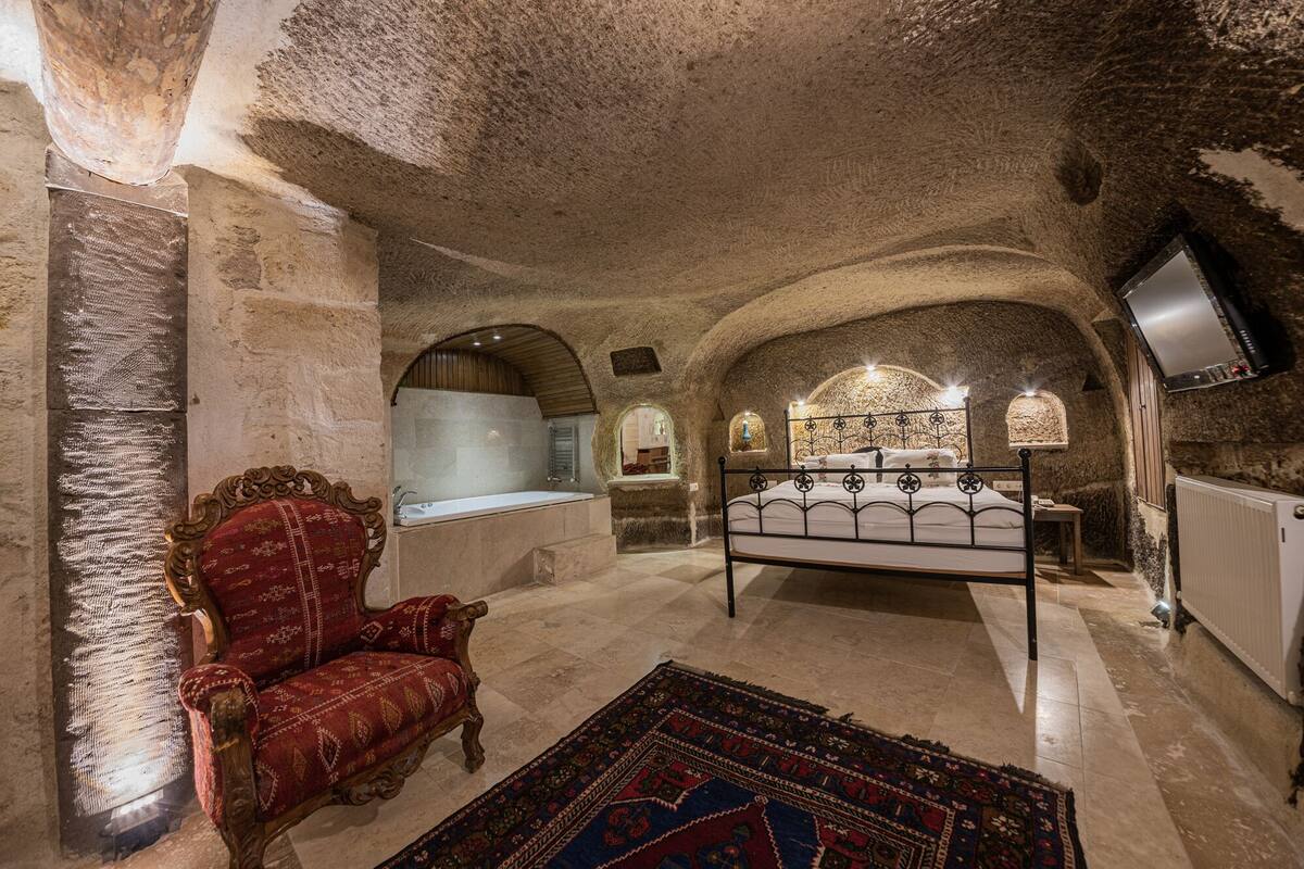 A room with a stone ceiling, a bed, a red upholstered chair, a small table, and a television mounted on the wall.
