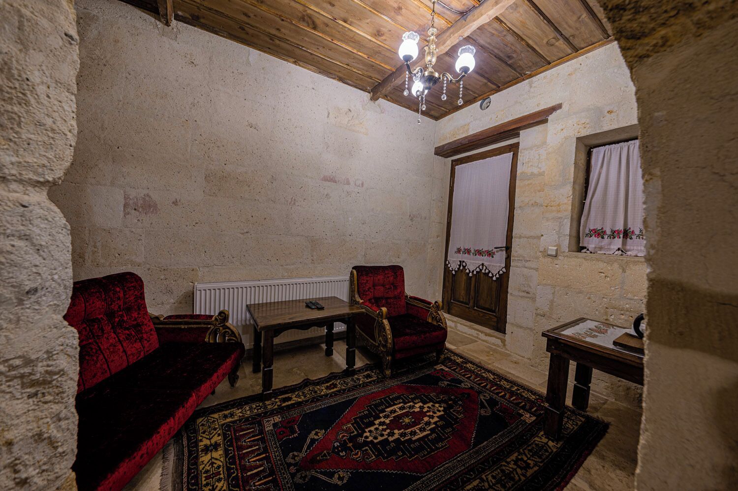 Una habitación rústica con techo de madera, un candelabro, un sofá de terciopelo rojo, una mesa de madera y un tapete estampado.
