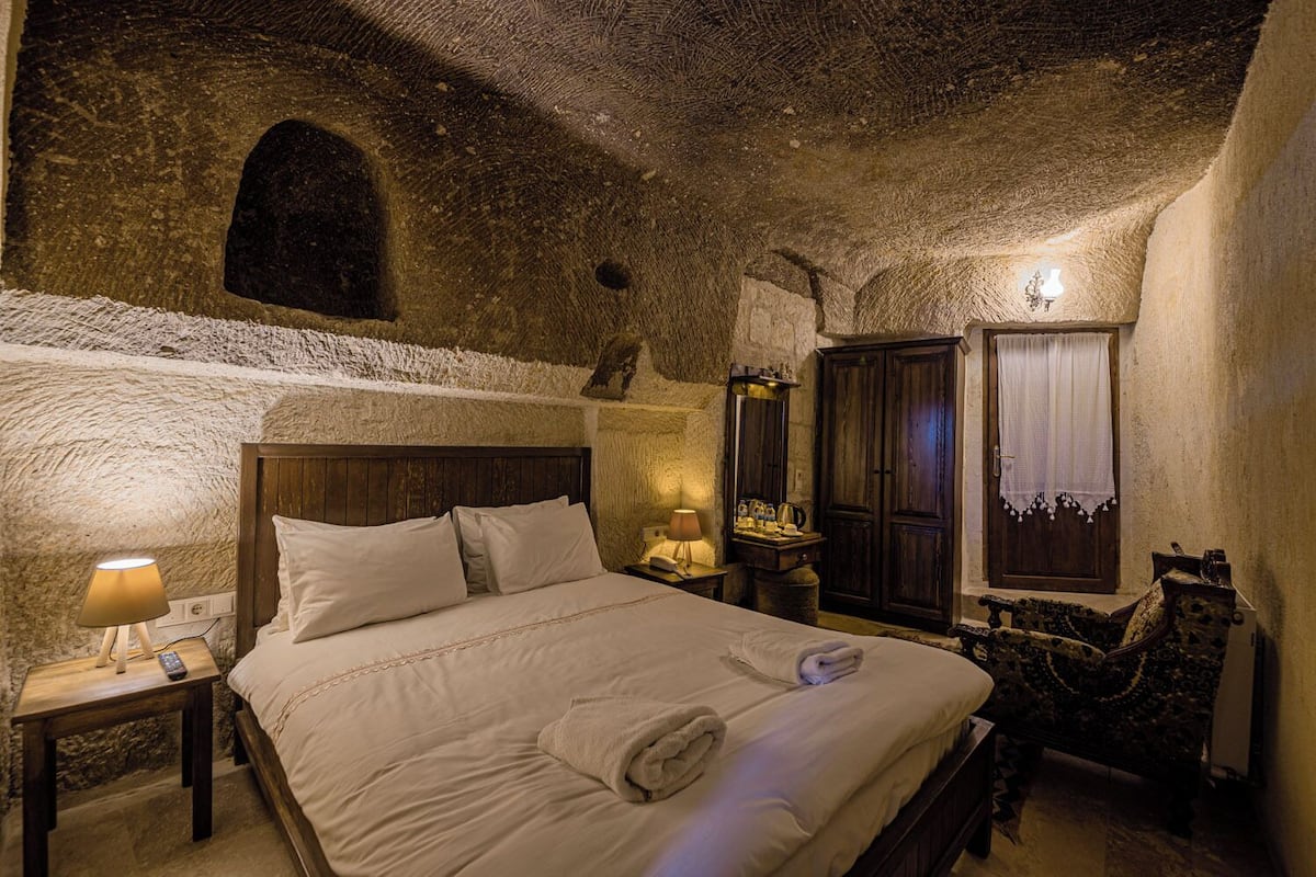 A cozy bedroom with a stone ceiling, a wooden bed, a nightstand with a lamp, a wardrobe, and a small table with a lamp.