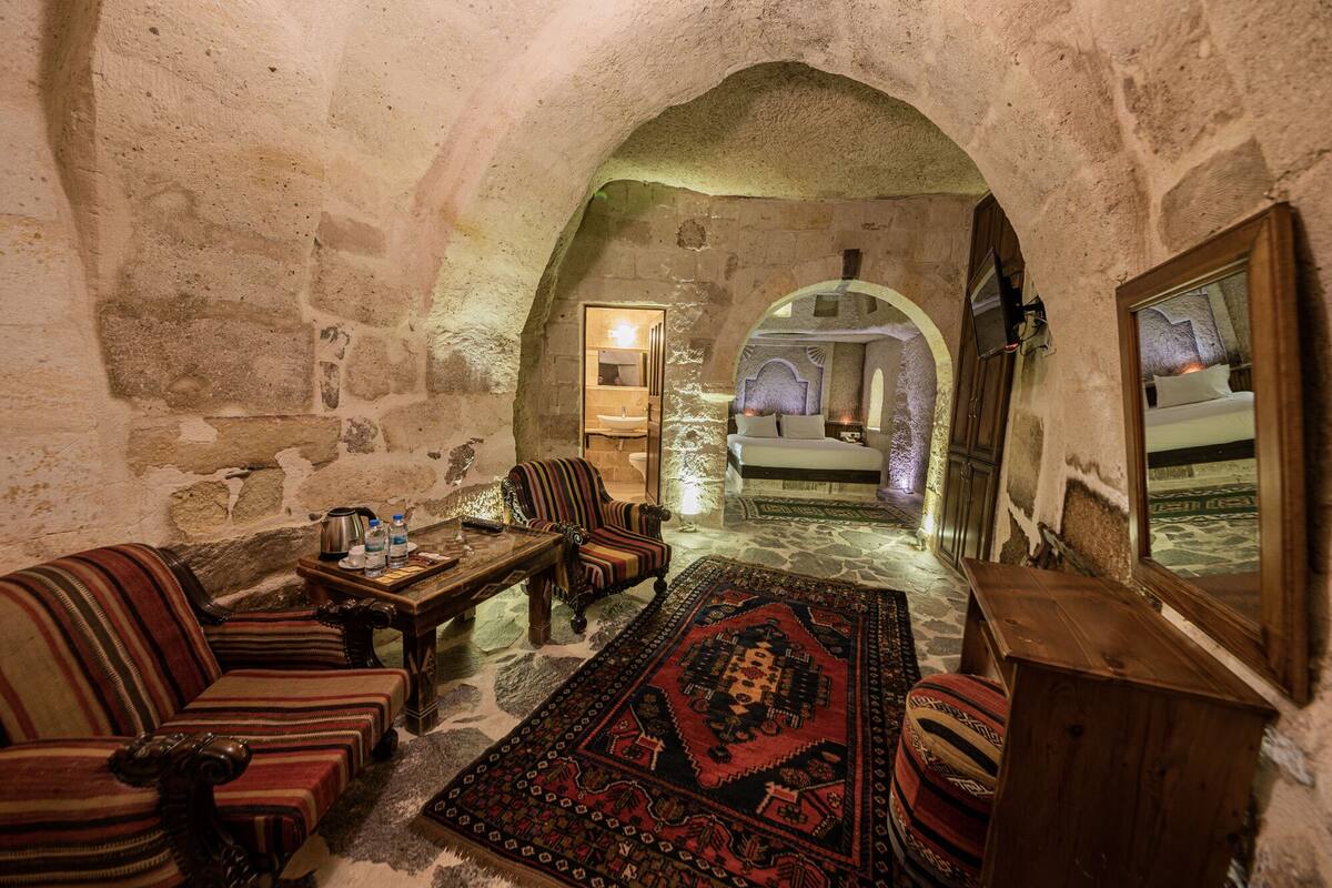 A cozy room with a stone wall, a wooden bed, a striped armchair, a small table with a kettle, and a rug.