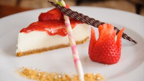 Una porción de pastel de queso con una decoración de fresa y chocolate sobre un plato blanco.