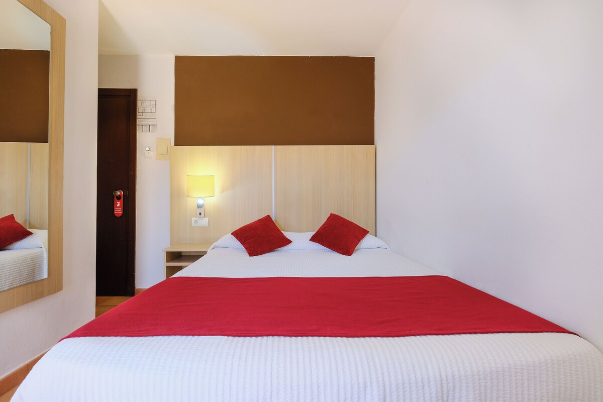 A hotel room with a large bed, red pillows, a wooden headboard, a nightstand with a lamp, and a mirror on the wall.