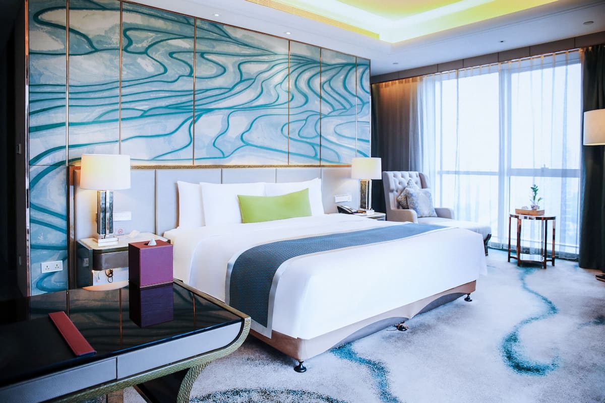 Ein modernes Hotelzimmer mit einem großen Bett, Nachttischlampen, einem Schreibtisch und Blick auf die Stadt durch große Fenster.