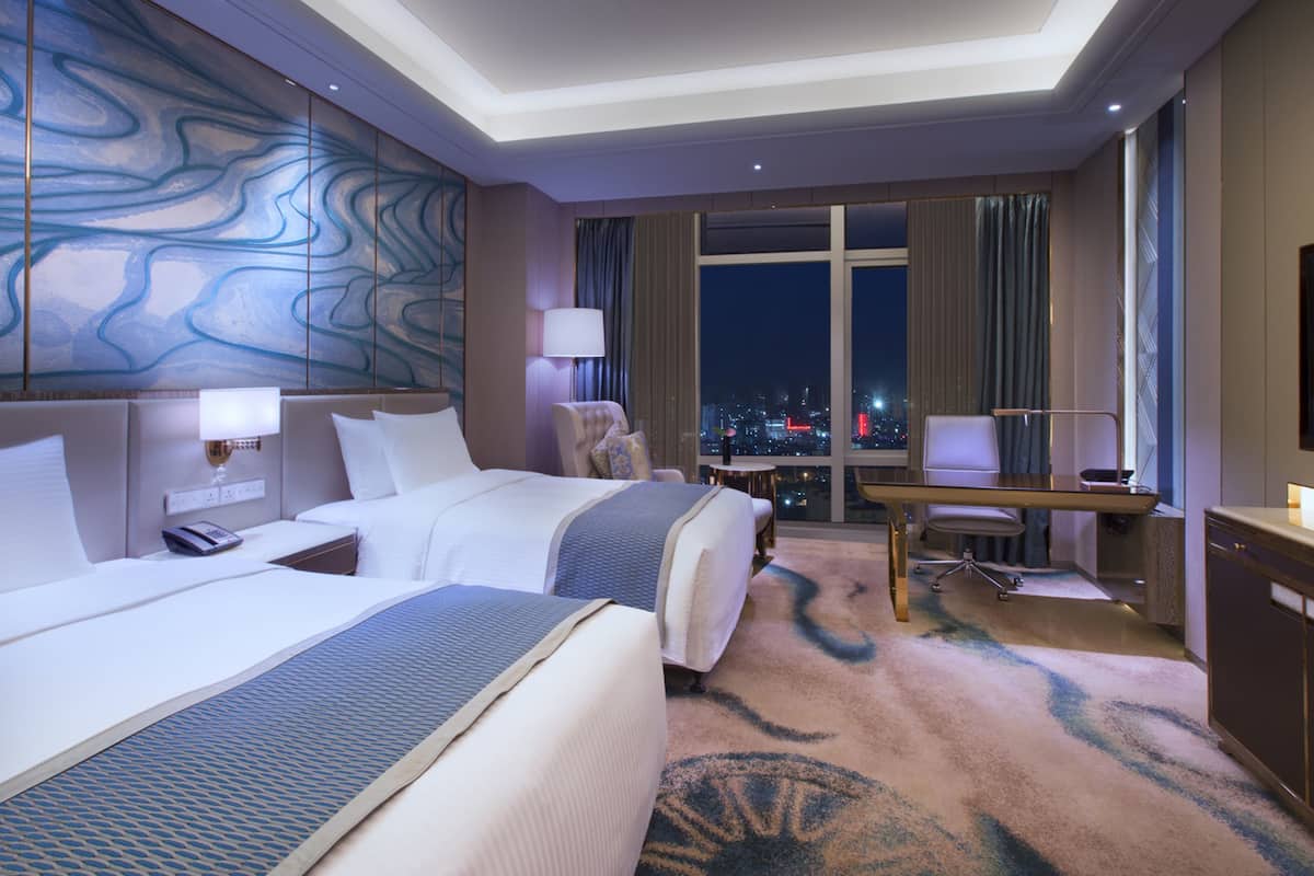 Ein Hotelzimmer mit zwei Betten, einem Schreibtisch, einem Stuhl und Blick auf die beleuchtete Stadt.