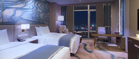 A hotel room with two beds, a desk, a chair, and a view of the city at night.