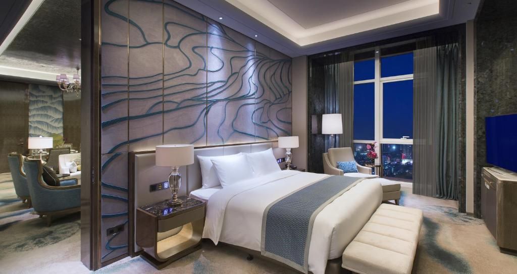 Una habitación de hotel moderna con una cama grande, mesitas de noche, un espacio para sentarse y vistas de la ciudad por la noche.