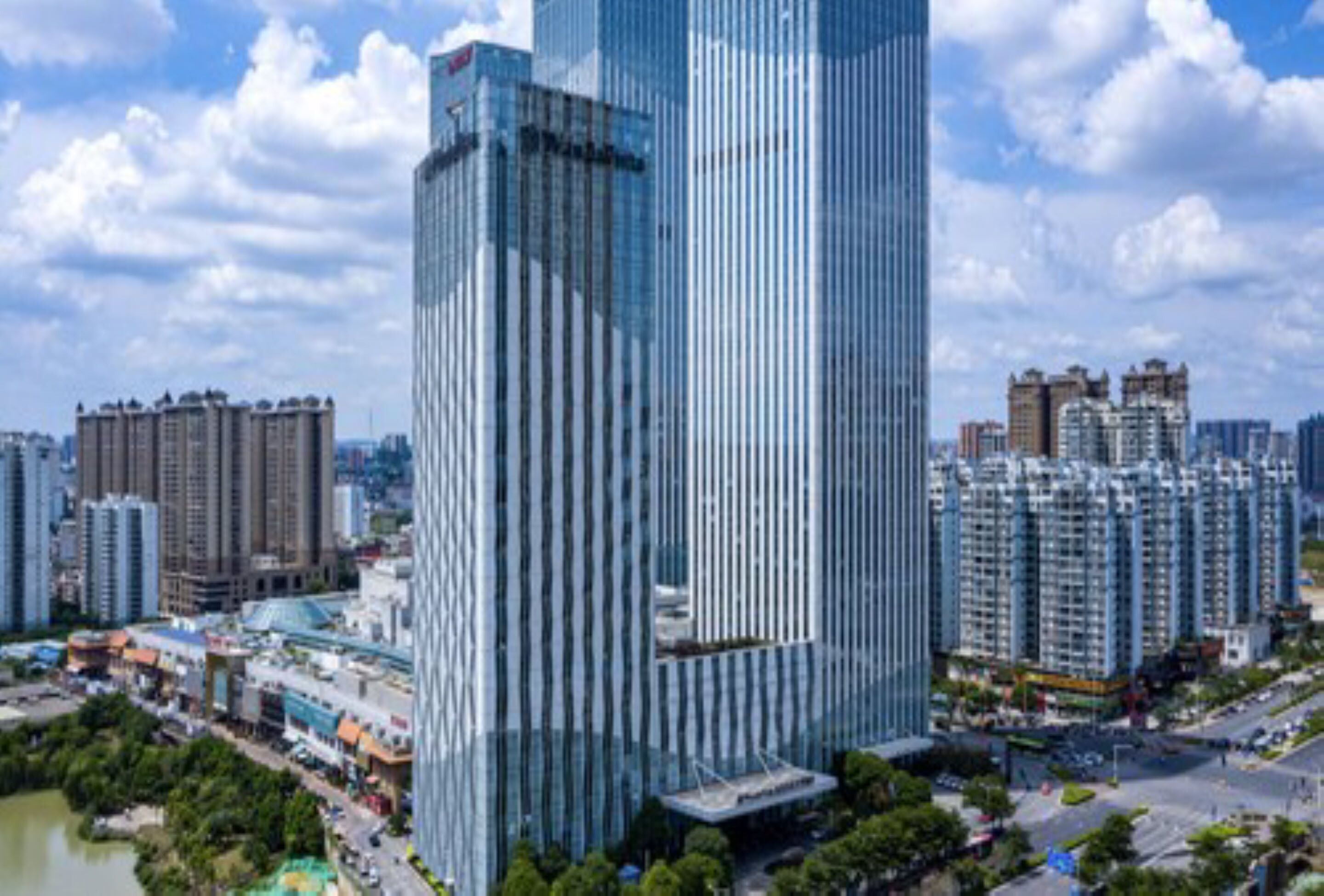 Un edificio de gran altura con fachada de vidrio moderna, rodeado de otros edificios residenciales y una masa de agua.