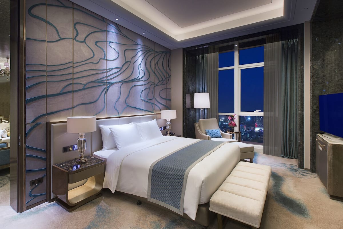 Ein modernes Hotelzimmer mit einem großen Bett, einer Sitzecke, Blick auf die beleuchtete Stadt und einer Wanddekoration.