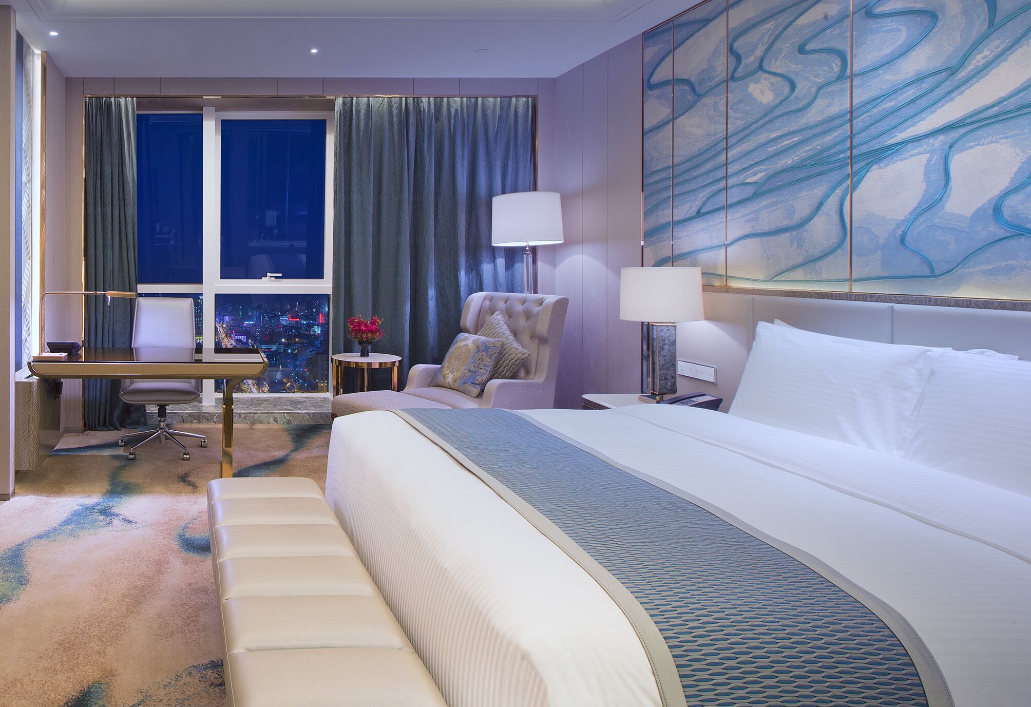 A hotel room with a large bed, a desk with a chair, a small table with a lamp, and a view of the city through the window.