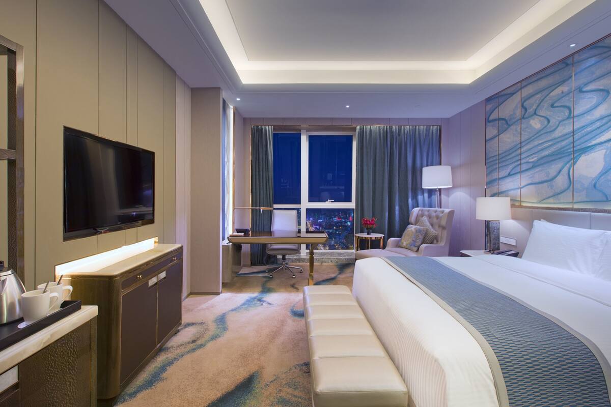 Ein Hotelzimmer mit einem großen Bett, einem Schreibtisch mit Stuhl, einem Fernseher und einem Fenster mit Blick auf die Stadt.