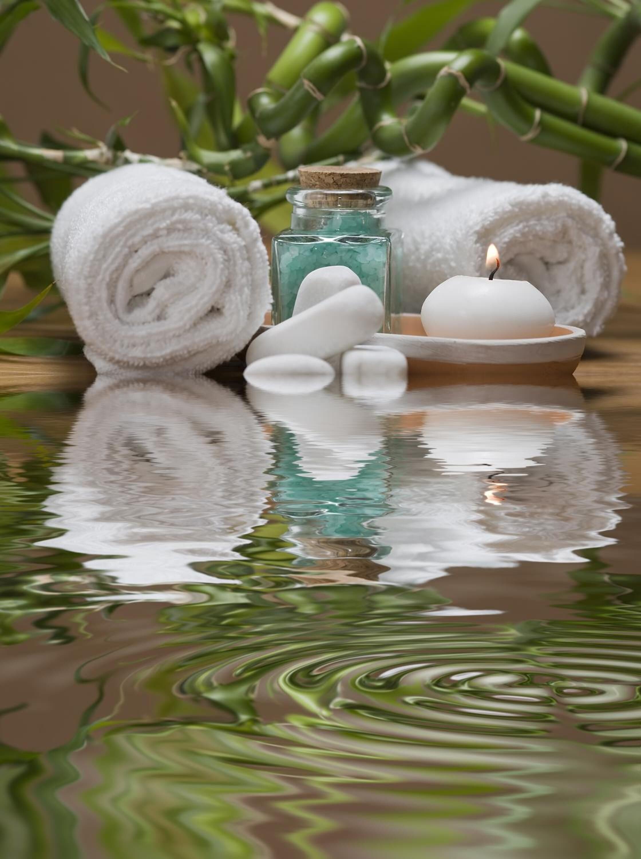 A spa setting with a candle, rolled towels, and a bottle of bath salts on a reflective surface.