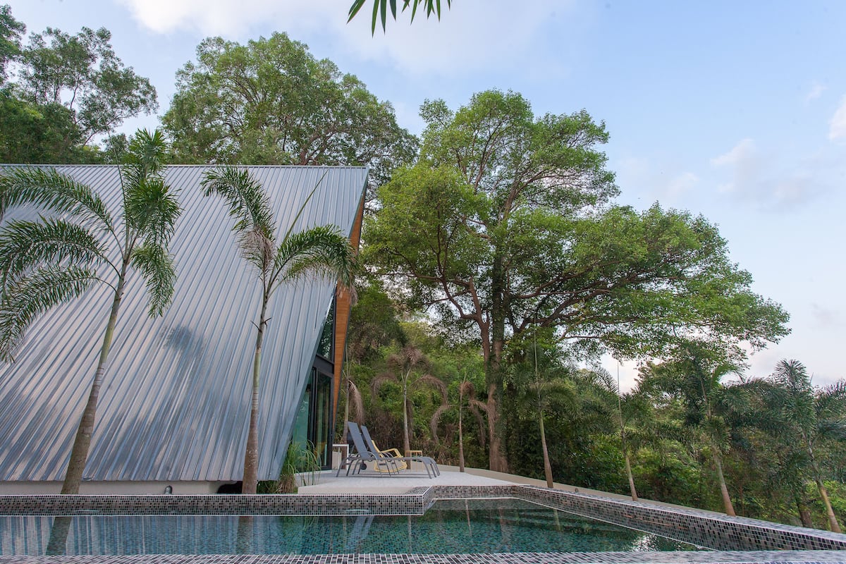 Un área de piscina moderna con una estructura de techo metálico, sillas de descanso y una piscina con bordes de azulejos.