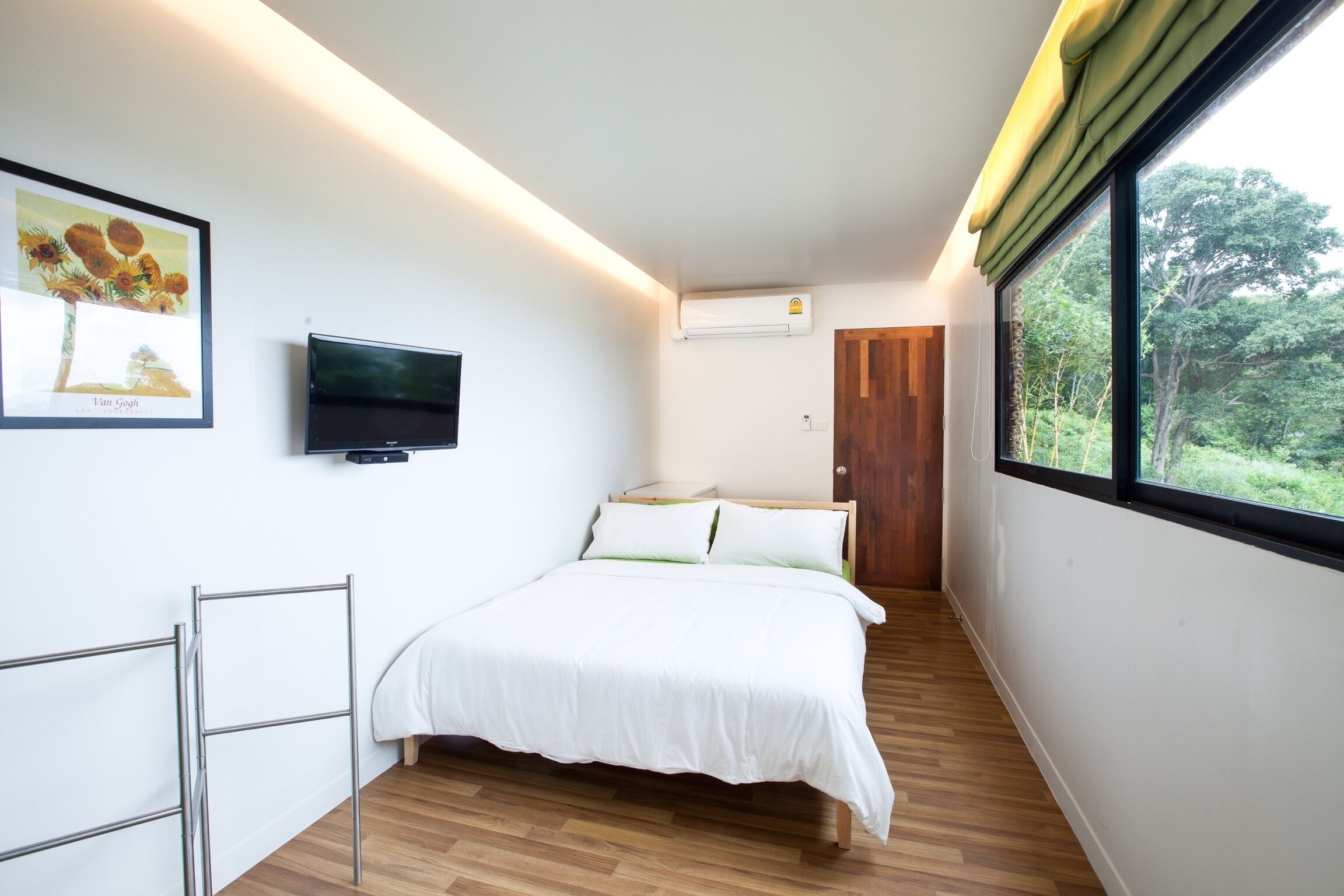 A bedroom with a bed, a TV, a window with a view of greenery, and a door.