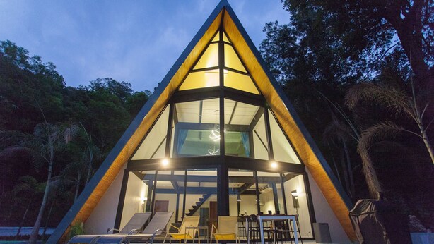Un edificio moderno de forma triangular con fachada de vidrio, una escalera que lleva a un nivel superior y un área de estar exterior con sillas y mesas.