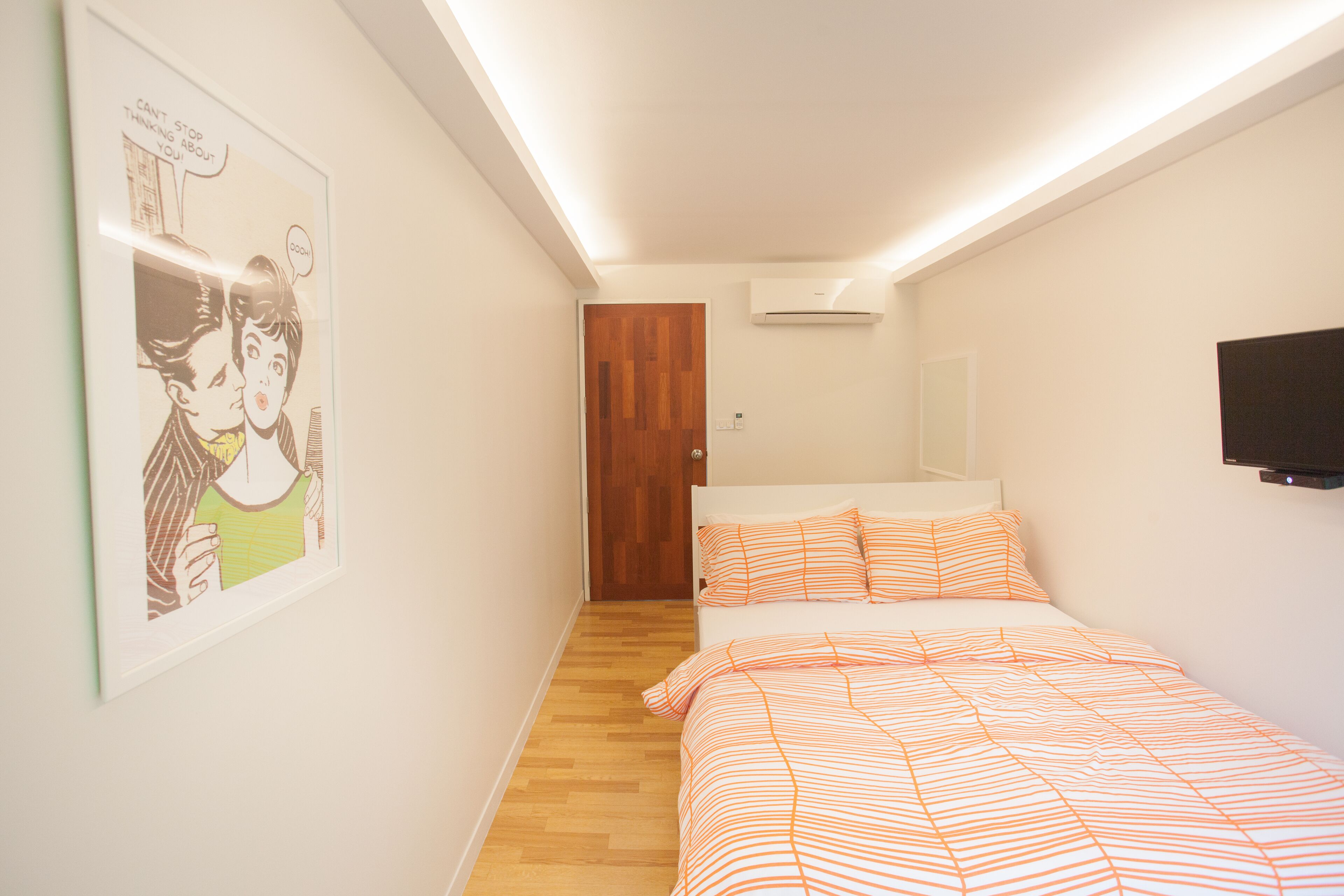 Ein kleines, helles Schlafzimmer mit Holztür, einem Bett mit gestreifter Bettwäsche, einem angebrachten Fernseher und einem Poster an der Wand.