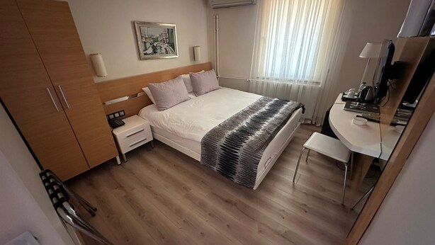 A hotel room with a wooden wardrobe, a bed with a patterned blanket, a bedside table with a lamp, a desk with a computer, and a window with curtains. - Sultan Hotel - Restaurant (Sivas)