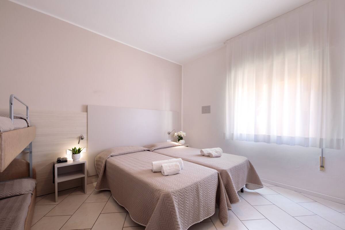 Une chambre d’hôtel avec deux lits, une petite table de chevet avec une lampe, un miroir et une fenêtre avec des rideaux légers.
