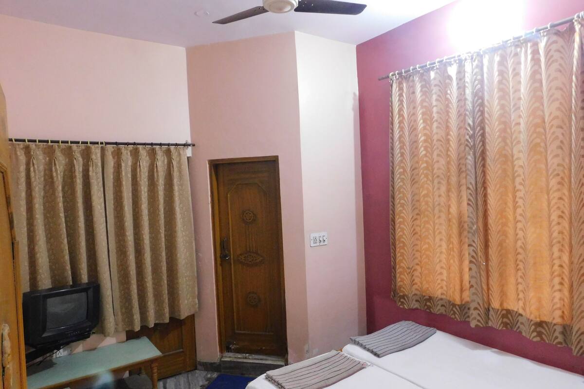 Une chambre avec un lit, un téléviseur posé sur un meuble, un ventilateur de plafond et des rideaux aux fenêtres.
