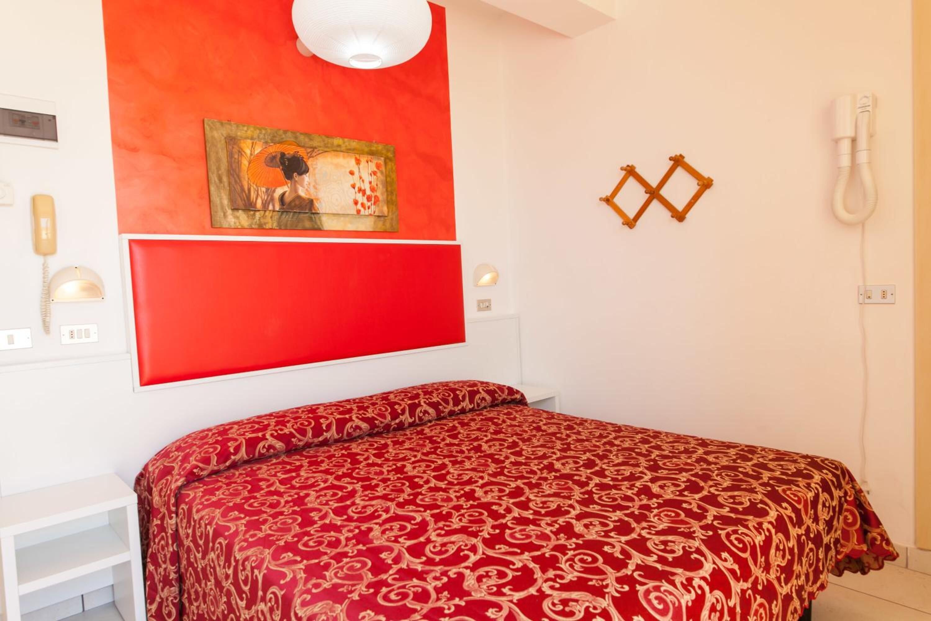 A bedroom with a red bedspread, a painting on the wall, and a telephone.