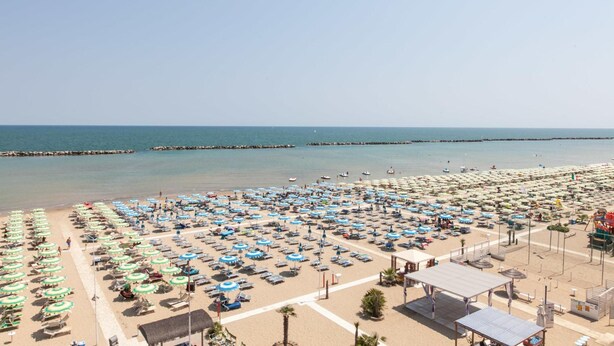 Ein Strand mit vielen Sonnenschirmen und Liegestühlen, ein Steg im Hintergrund und ein klarer Himmel.
