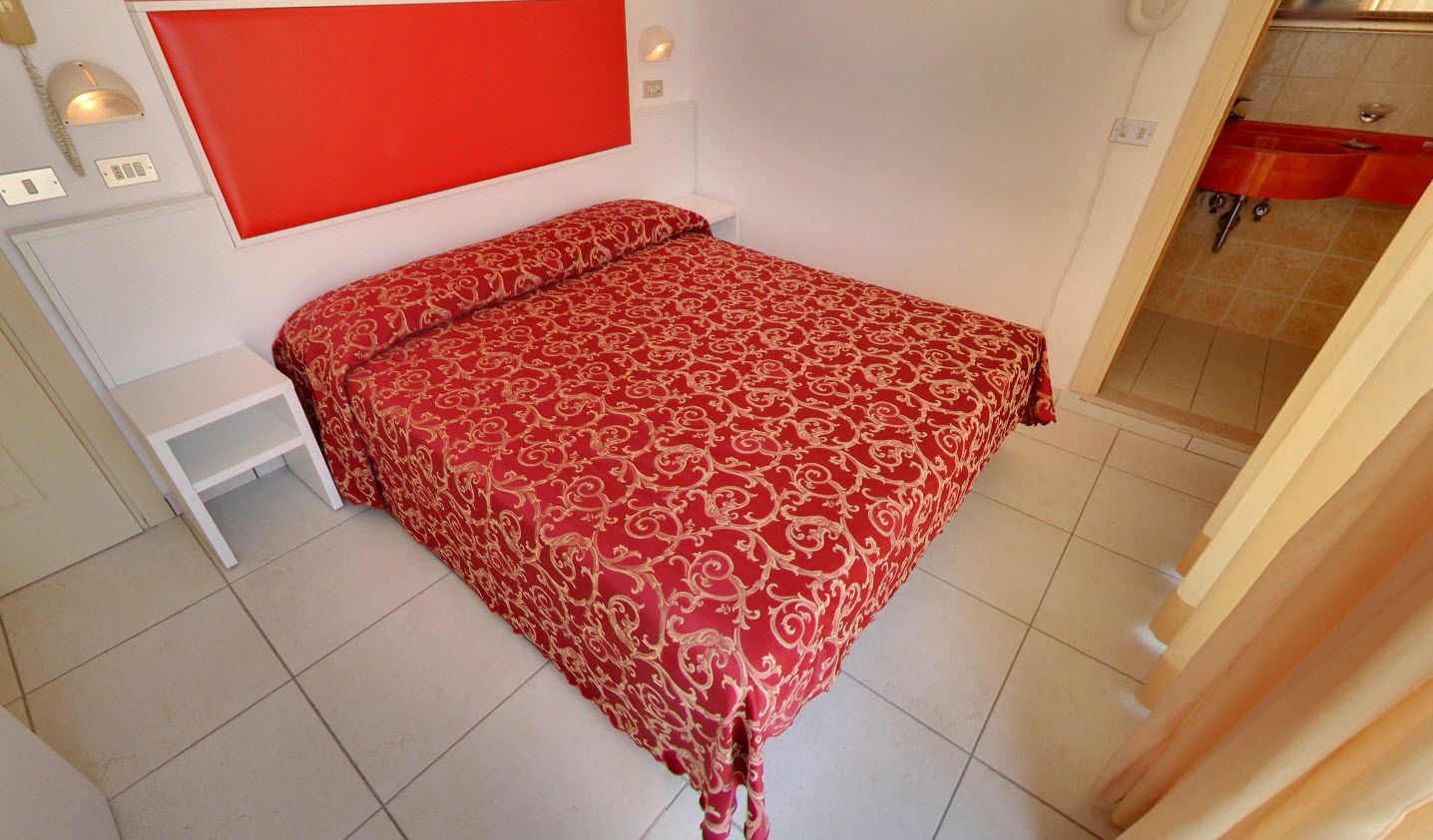 Un dormitori amb un llit, una tauleta de nit, un panell de capçal vermell i un bany visible a través d'una porta oberta.