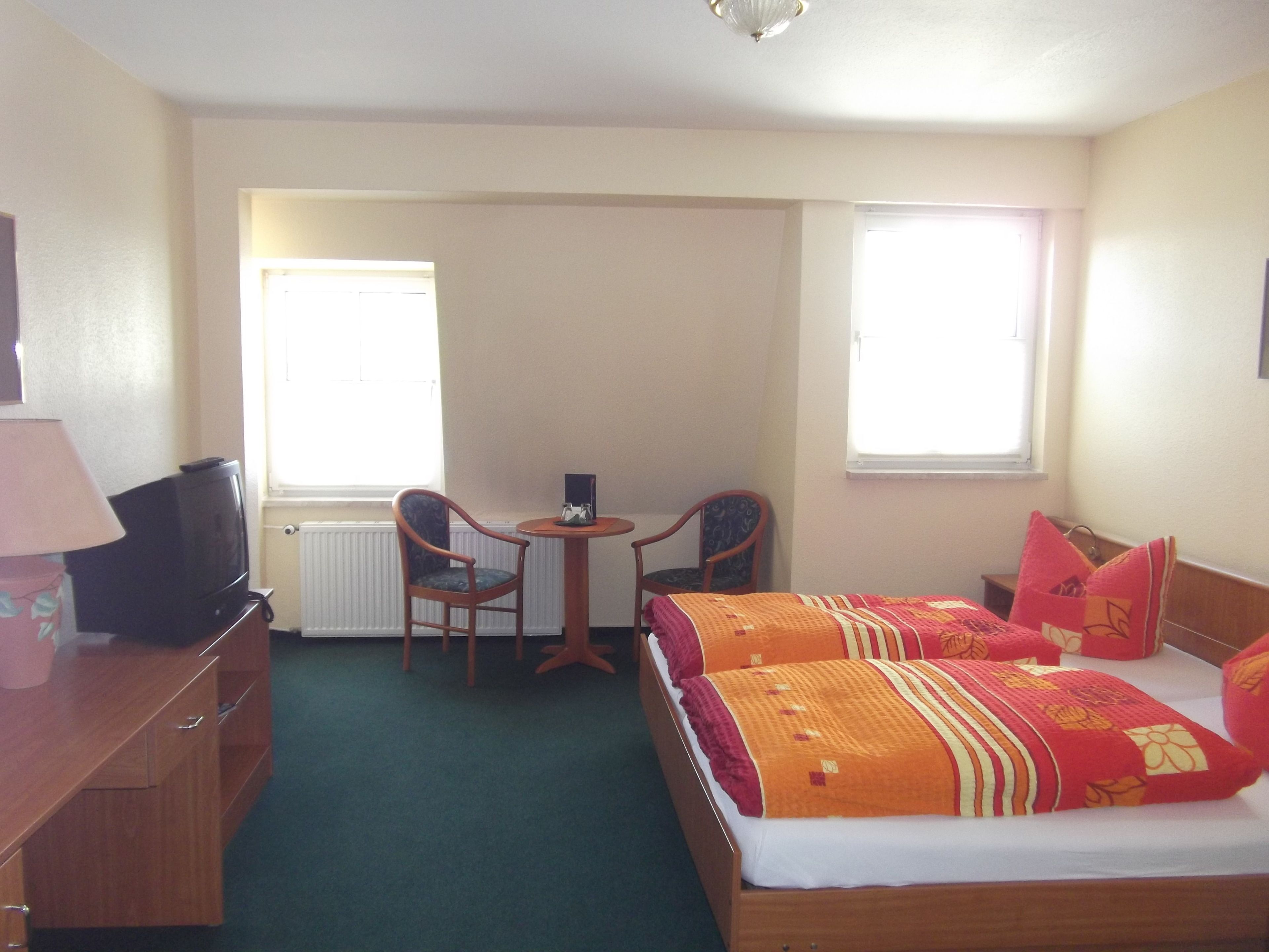 A hotel room with a bed, a desk with a lamp, two chairs, and a television.