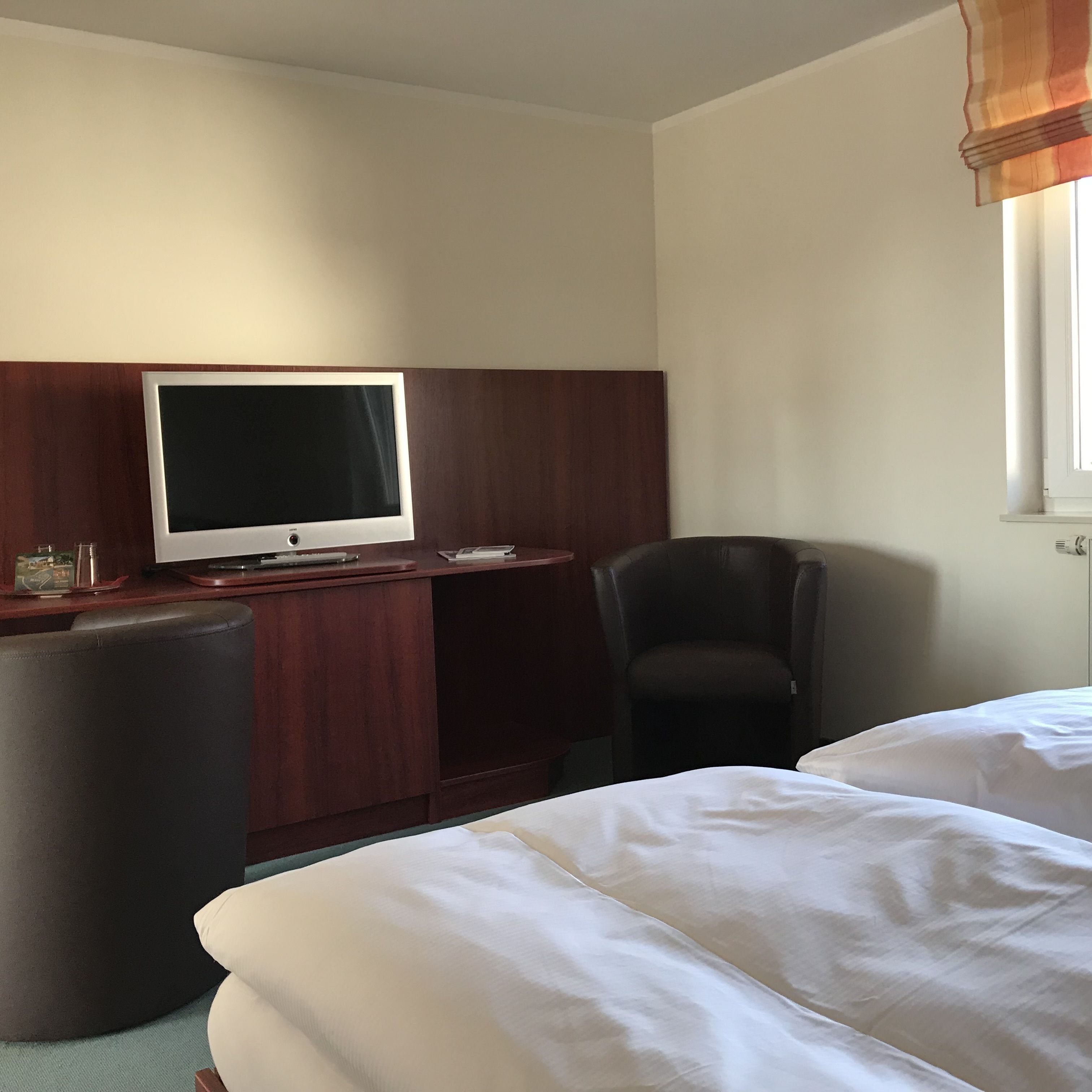 A hotel room with a wooden desk, a flat-screen TV, a black chair, and two beds with white linens.