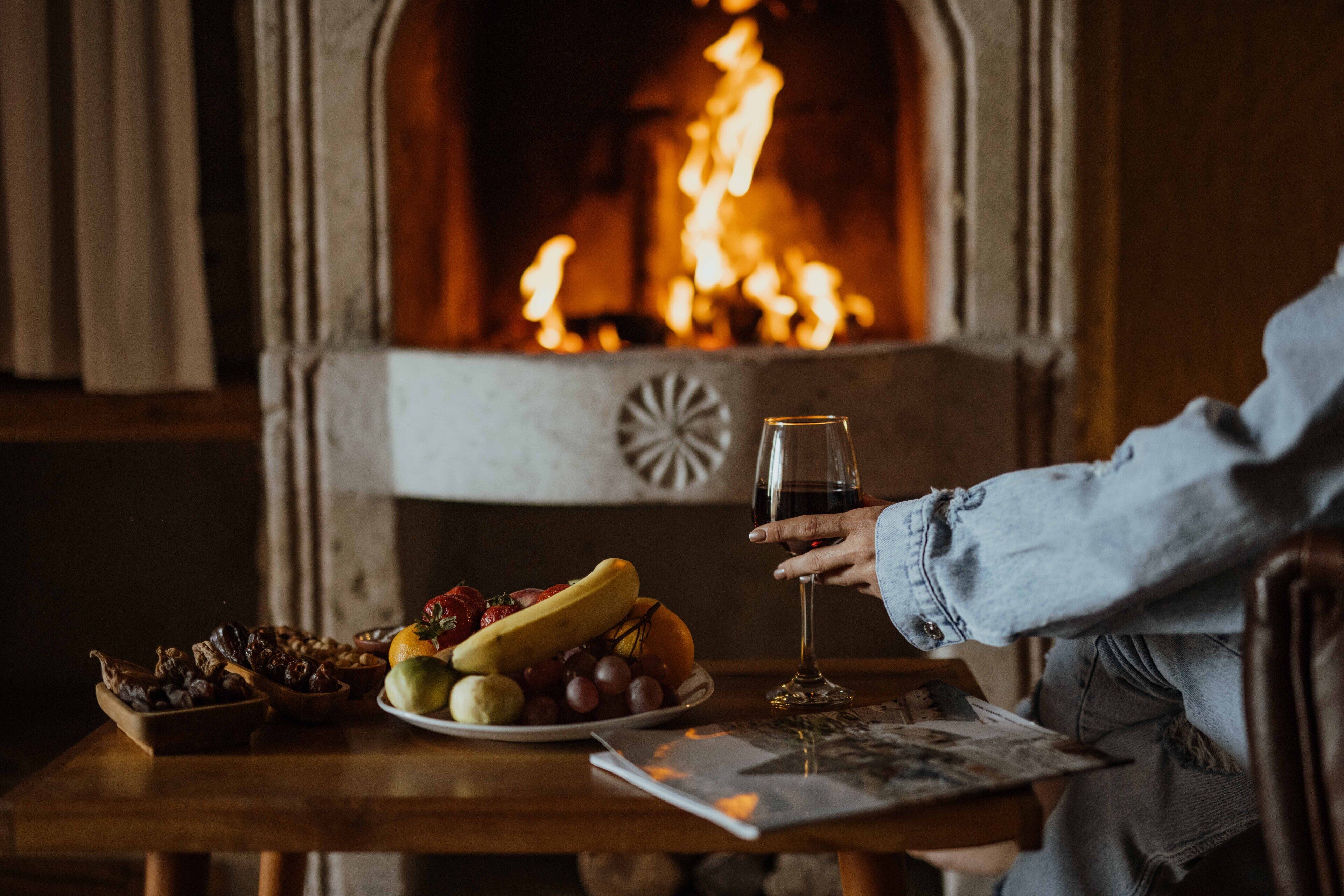 Uma pessoa segurando um copo de vinho em uma mesa de madeira com um prato de frutas e um jornal. Uma lareira com manto de pedra está ao fundo.